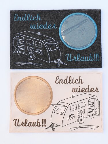 Zwei gestickte Tassenteppiche mit Wohnwagen-Motiv und Schrift 'Endlich wieder Urlaub!!!', oben dunkelgrau mit türkis, unten hellbeige mit braunen Stickereien