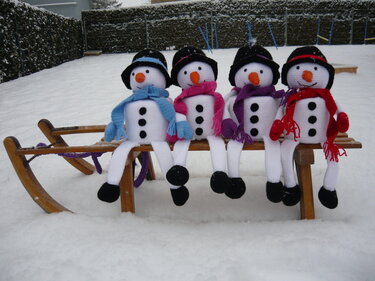 Vier gehäkelte Schneemänner mit schwarzen Hüten und bunten Schals auf einem Holzschlitten im Schnee
