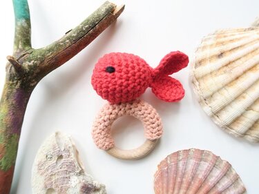 Gehäkelte rote Fisch-Rassel auf hellem Holz-Beißring, umgeben von Muscheln und Treibholz
