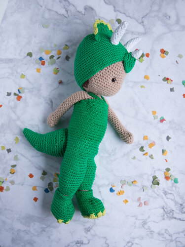 Gehäkelte Amigurumi-Puppe im grünen Triceratops-Kostüm mit Kapuze, Hörnern und Schwanz auf marmoriertem Hintergrund mit Konfetti.