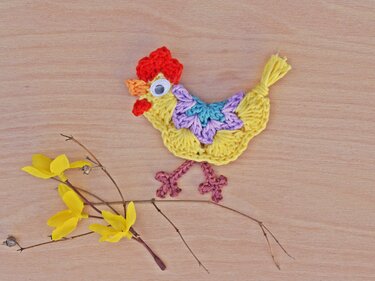 Gehäkelte gelbe Küken-Applikation mit buntem Flügelmuster und Wackelauge auf Holz neben gelben Zweigen