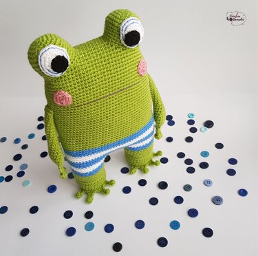 Grüne gehäkelte Amigurumi-Froschfigur mit großen Augen, rosigen Wangen und blau-weiß gestreifter Hose auf hellem Untergrund mit verstreuten blauen Knöpfen