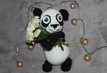 Black and white crocheted panda holding white chrysanthemums with warm string lights on a grey background.