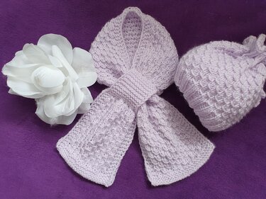 Helllila gestrickte Babymütze und passender Schal mit Strukturmuster auf violettem Untergrund, daneben eine weiße Stoffblüte.