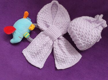 Gestrickte Babymütze und passender Schal in hellem Flieder mit Strukturmuster, daneben blaues Elefanten‑Greifspielzeug auf violettem Untergrund.