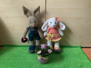Zwei gehäkelte Amigurumi-Osterhasen: grauer Hase mit blauer Latzhose und Körbchen; weißes Häschen im rosa Kleid mit Blüte