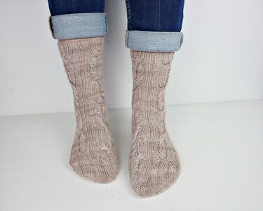 Beige Zopfmustersocken an Damenbeinen mit hochgekrempelten Jeans vor hellem Hintergrund.
