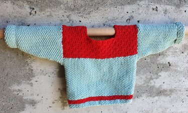 Baby-Pullover in Hellblau mit rotem Einsatz und Saumstreifen, gestrickt im Krausmuster, an einer Holzstange präsentiert.