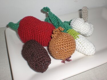 Fünf gehäkelte Gemüsefiguren: Paprika, Zwiebel, Lauch, Kartoffel und Pilz auf weißer Platte.