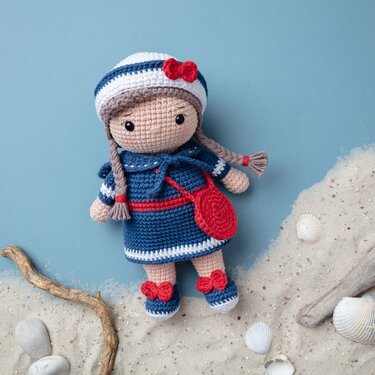 Gehäkelte Amigurumi-Puppe mit blauem Kleid, maritimer Mütze mit roter Schleife und roter Tasche auf sandigem Hintergrund mit Muscheln