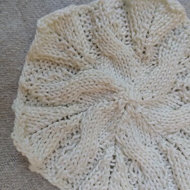 Gestrickte cremefarbene Mütze mit spiralförmigen Zopf- und Ajourreihen auf grauem Untergrund