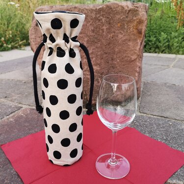 Beigefarbene Stoff‑Flaschentasche mit schwarzen Tupfen und schwarzem Kordelzug neben leerem Weinglas auf rotem Tuch