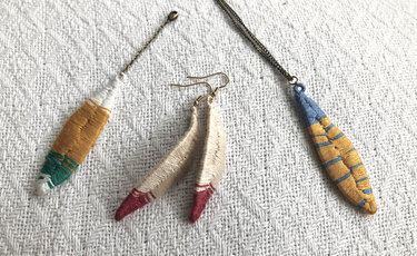 Zwei bestickte Anhänger und ein Paar bestickte Hängeohrringe auf weißem, strukturiertem Stoff; Farben: Senfgelb, Blau, Grün, Beige, Rot.