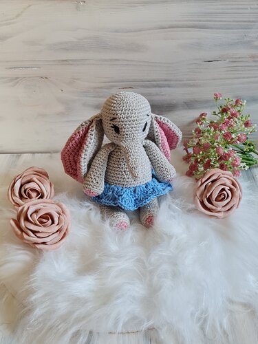 Grauer gehäkelter Amigurumi-Elefant mit rosa Ohren und blauem Röckchen, sitzend auf weißem flauschigem Untergrund mit Rosenblüten.