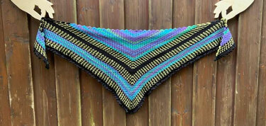 Gehäkeltes Dreieckstuch mit V‑förmigen Streifen in Schwarz, Gelb, Blau, Türkis und Lila vor Holzlatten