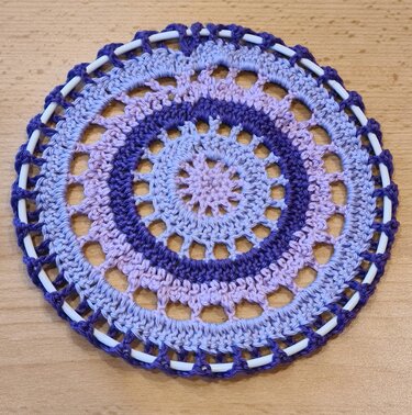 Nahaufnahme eines runden gehäkelten Mandalas in Lila, Lavendel und Hellblau um einen weißen Ring auf einer Holzoberfläche.