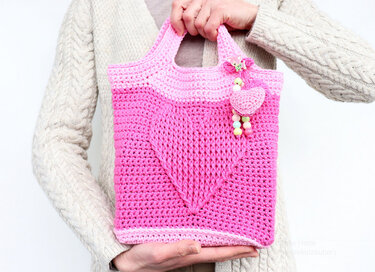 Hand hält pinke gehäkelte Henkeltasche mit eingearbeitetem Herz und Perlenanhänger