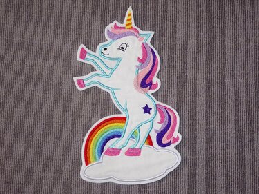 Weißes Einhorn-Stickmotiv mit rosa-violetter Mähne und Schweif, gelbem Horn, pinken Hufen und kleinem lila Stern auf der Flanke, auf einer weißen Wolke vor einem Regenbogen.