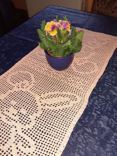 Cremefarbener gehäkelter Tischläufer mit Rauten- und Lochmuster auf dunkelblauem Tischtuch, dekoriert mit einem kleinen Blumentopf.