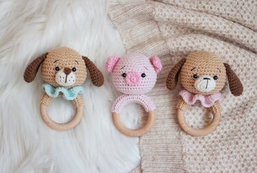 Three crochet baby rattles: two brown dogs with blue and pink ruffles, and one pink pig, all with wooden rings, on a fluffy white and knitted beige background.