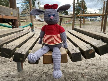 Gehäkelter grauer Amigurumi-Hase mit rotem Pullover und Schleife sitzt auf einer Holzbrücke