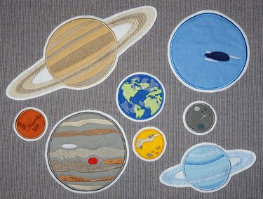 Acht bestickte Planetentextil-Patches (z. B. Saturn, Jupiter, Erde, Mars, Mond) auf grauem Stoffhintergrund.