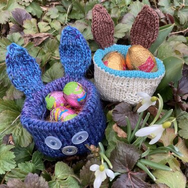 Zwei gestrickte Körbchen in Hasenform in Blau und Natur, mit Ohren und Knopfaugen, gefüllt mit Schokoeiern zwischen Blättern und Schneeglöckchen