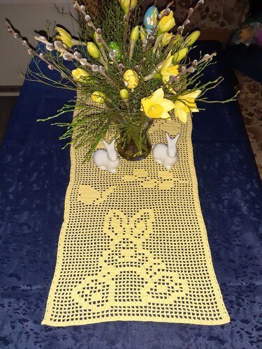 Gehäkelter gelber Tischläufer mit Narzissen, Weidenkätzchen und zwei weißen Hasenfiguren auf blauem Tisch