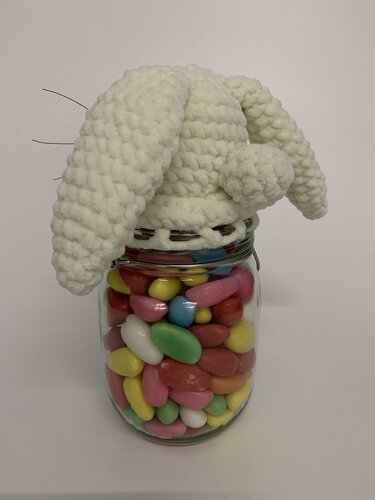 Ein Einmachglas voller pastellfarbener Dragees, bedeckt mit einer abnehmbaren weißen gehäkelten Hasenmütze mit Schlappohren