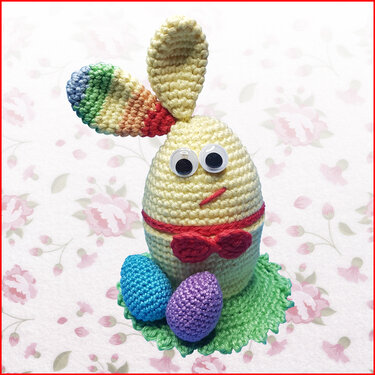 Gehäkelter Eierwärmer als Hase mit Kulleraugen, roter Fliege, regenbogenfarbenem Ohr und zwei gehäkelten Ostereiern auf grünem Untersetzer.