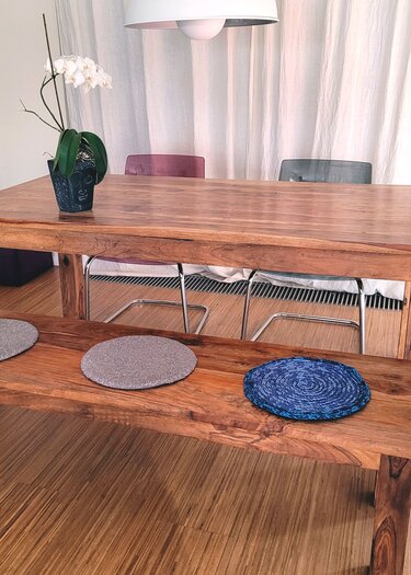 Essbereich mit Holztisch, Holzbank und drei runden Tischsets in Grau und Blau; weiße Orchidee in einem Topf mit Buddha-Motiv auf dem Tisch.