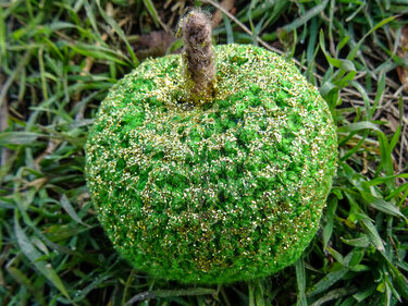 Gehäkelter grüner Apfel mit goldfarbenem Glitzer und braunem Stiel auf Gras