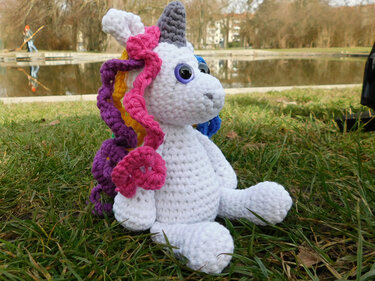 Weißes gehäkeltes Einhorn-Amigurumi mit Regenbogenmähne und grauem Horn auf Gras neben einem Teich.