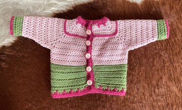 Gehäkelte Baby-Cardigan in Rosa und Grün mit Fuchsia-Rändern und fünf hellen Knöpfen auf braunem Hintergrund