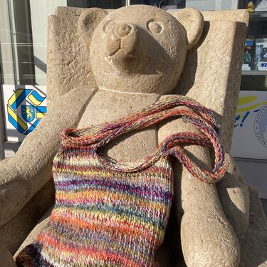 Multicolored knitted shoulder bag with long strap draped on a carved stone bear bench