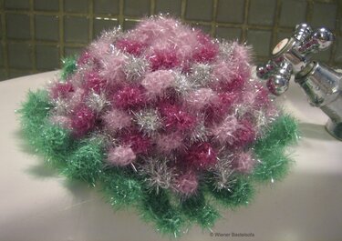 Nahaufnahme eines runden Häkelpads aus Effektgarn mit grünem Rand und rosa-silbernen fluffigen Maschen am Waschbeckenrand