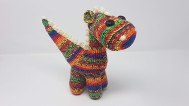 Amigurumi-Dinosaurier in Regenbogenstreifen mit weißen Zacken und schwarzen Augen auf weißem Hintergrund.