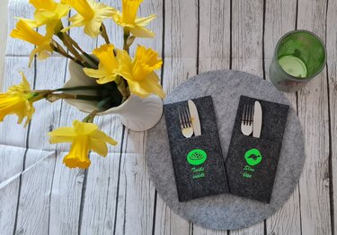 Zwei dunkelgraue Filz-Bestecktaschen mit neongrünen Schildkröten-Plotmotiven und englischen Schriftzügen, auf rundem Filzuntersetzer platziert; links weiße Vase mit gelben Narzissen und rechts ein grünes Glas