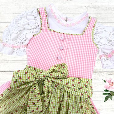 Kinder-Dirndl mit rosa-kariertem Oberteil, grüner Blümchenschürze und weißer Spitzenbluse auf hellem Holzuntergrund