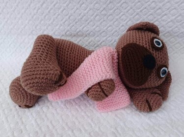Gehäkelter brauner Amigurumi Hund mit rosa Schal auf weißem Untergrund liegend