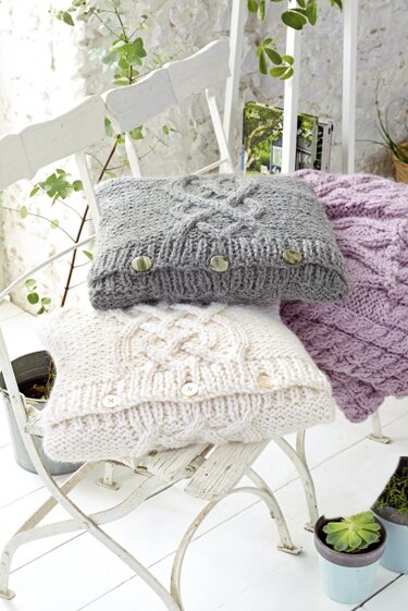 Drei gestrickte Kissenhüllen in Grau, Creme und Flieder mit Zopfmuster und Knöpfen auf weißen Stühlen