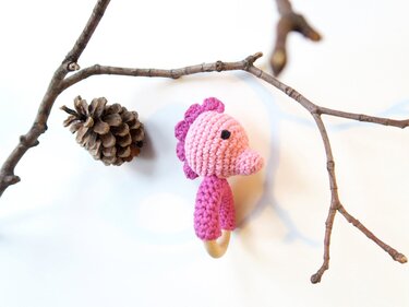 Rosa gehäkeltes Seepferdchen an Holzring neben Zweigen und Zapfen