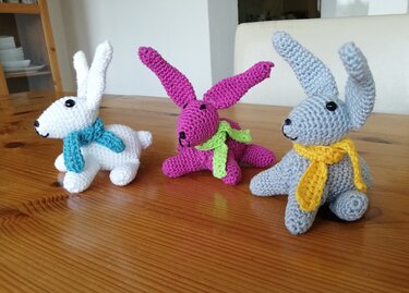 Drei gehäkelte Amigurumi-Hasen in Weiß, Pink und Grau mit bunten Schals auf Holztisch