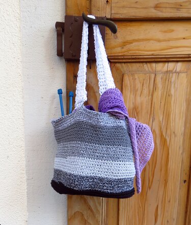 Gehäkelte Henkeltasche mit grauen und weißen Streifen, aus der blaue Stricknadeln und lila Garn ragen, an einer Holztür hängend.