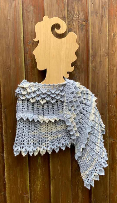 Hellblaues gehäkeltes Tuch mit Drachenschuppenrand, drapiert auf einer hölzernen Büste vor einer Holzwand.