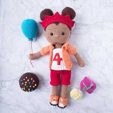 Gehäkelte Amigurumi-Puppe mit brauner Haut und braunen Haaren, roter Krone, Shirt mit roter '4', roter Hose und orangen Schuhen. Sie hält einen hellblauen Ballon; daneben gehäkelter Kuchen, Geschenk und Garnknäuel.