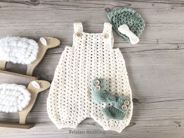 Gehäkelter beigefarbener Strampler, mintgrüne Mütze mit Schleife und mintgrüne Babyschühchen auf hellem Holzhintergrund neben Holzschafen.