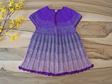 Gehäkeltes Babykleid in Lila-Farbverlauf liegend auf Holz mit gelben Blütenzweigen