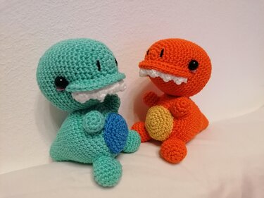 Zwei gehäkelte Amigurumi-Dinos in Mint und Orange mit weißen Zähnchen und schwarzen Sicherheitsaugen