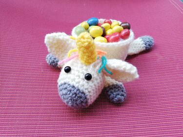 Handgehäkelter weißer Einhorn-Korb mit gelbem Horn, bunter Mähne und grauen Akzenten, gefüllt mit bunten Bonbons auf pinkem Untergrund.